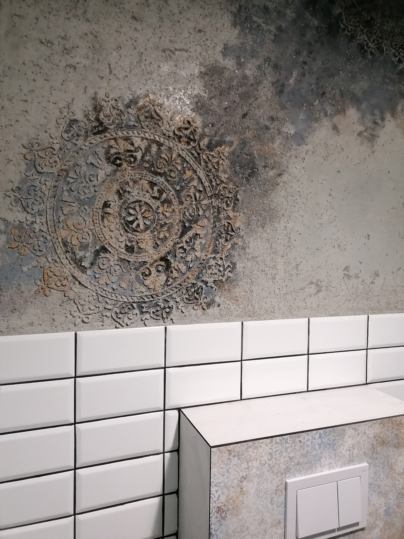 Dekoracyjna szpachla z motywem mandali na ścianie nad białymi, prostokątnymi płytkami w łazience z zabudowaną spłuczką.