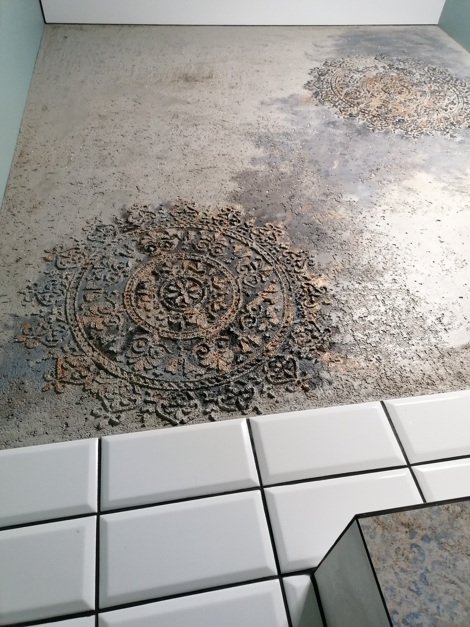 Ściana z szarą, teksturowaną farbą imitującą beton, z dekoracyjnymi, okrągłymi wzorami przypominającymi ornamenty, wykończona białymi, prostokątnymi płytkami typu metro z ciemną fugą.