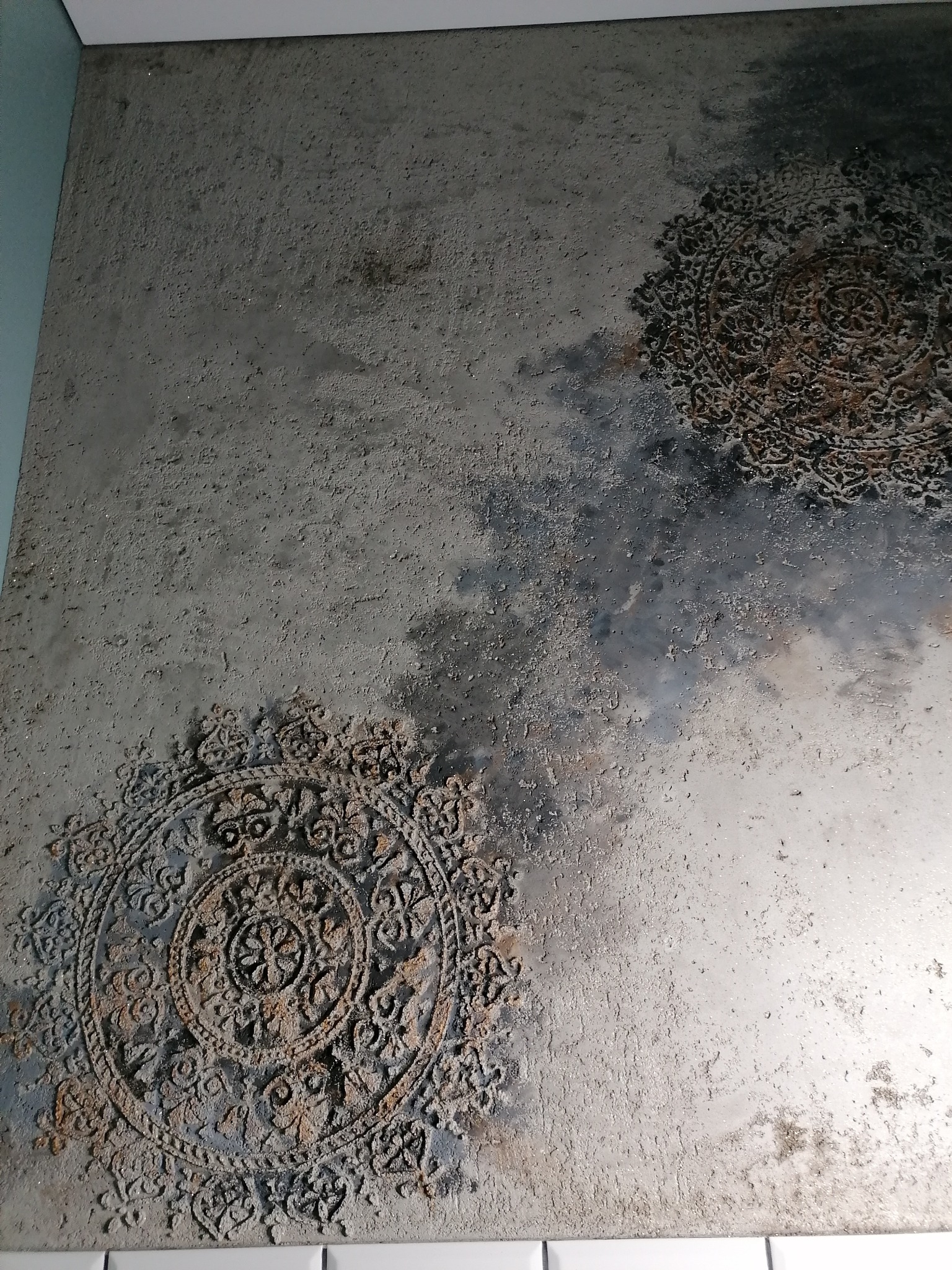 Dekoracyjna ściana z efektem betonu i wzorami mandali w odcieniach szarości i brązu, z widoczną strukturą i nierównościami.