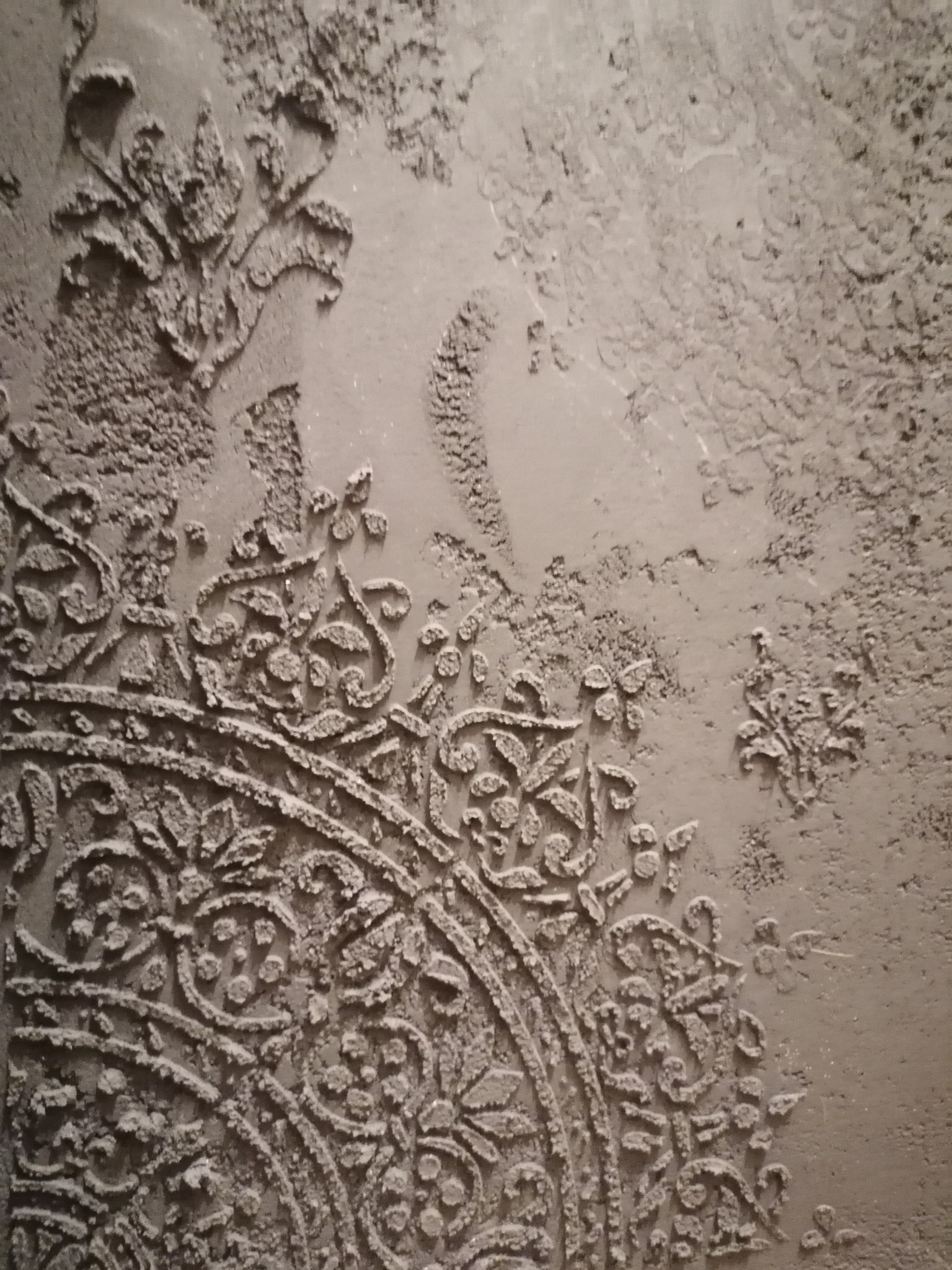 Dekoracyjna ściana z szarą, teksturowaną powierzchnią i wypukłym, kwiatowym wzorem w stylu vintage, wykonana techniką szpachlowania.