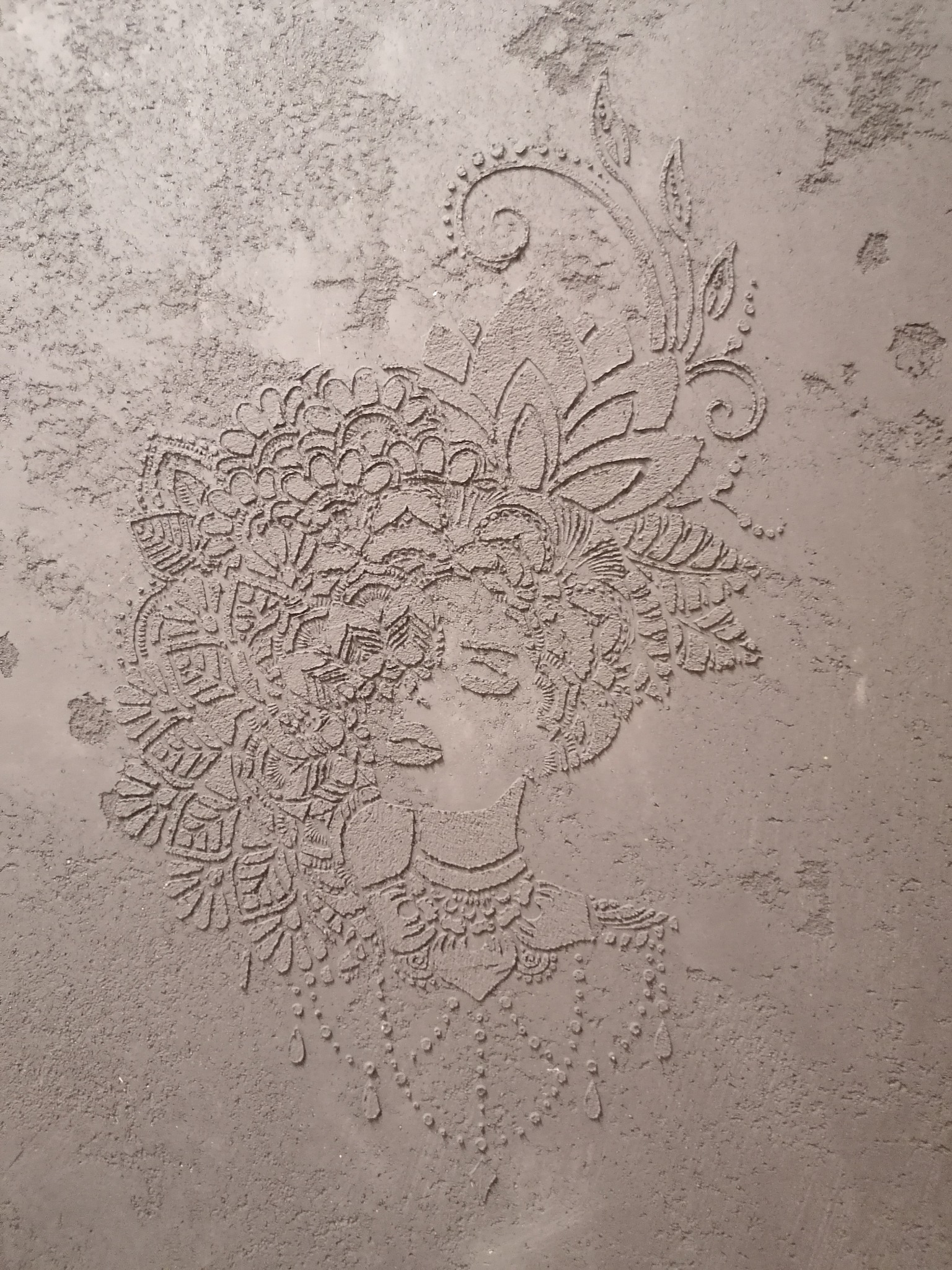 Dekoracyjna struktura na ścianie przedstawiająca profil kobiety w bogatym nakryciu głowy, wykonana w odcieniach szarości, z widocznymi nierównościami i fakturą.