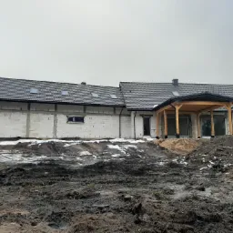 Surowy stan domu jednorodzinnego w trakcie budowy, widoczny szary dach z oknami połaciowymi, białe ściany z bloczków, drewniana konstrukcja zadaszenia tarasu, błotniste podłoże z resztkami śniegu.