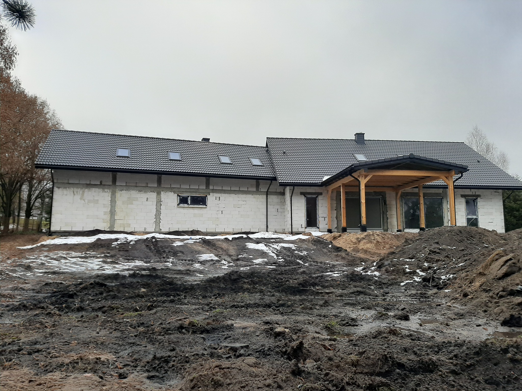 Surowy stan domu jednorodzinnego w trakcie budowy, widoczny szary dach z oknami połaciowymi, białe ściany z bloczków, drewniana konstrukcja zadaszenia tarasu, błotniste podłoże z resztkami śniegu.