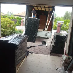 Mężczyzna w trakcie załadunku mebli owiniętych czarną folią do wnętrza samochodu dostawczego, widok z wnętrza garażu na posesję z domem i zielenią.