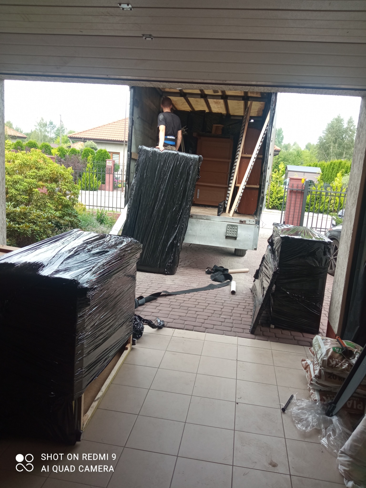 Mężczyzna w trakcie załadunku mebli owiniętych czarną folią do wnętrza samochodu dostawczego, widok z wnętrza garażu na posesję z domem i zielenią.