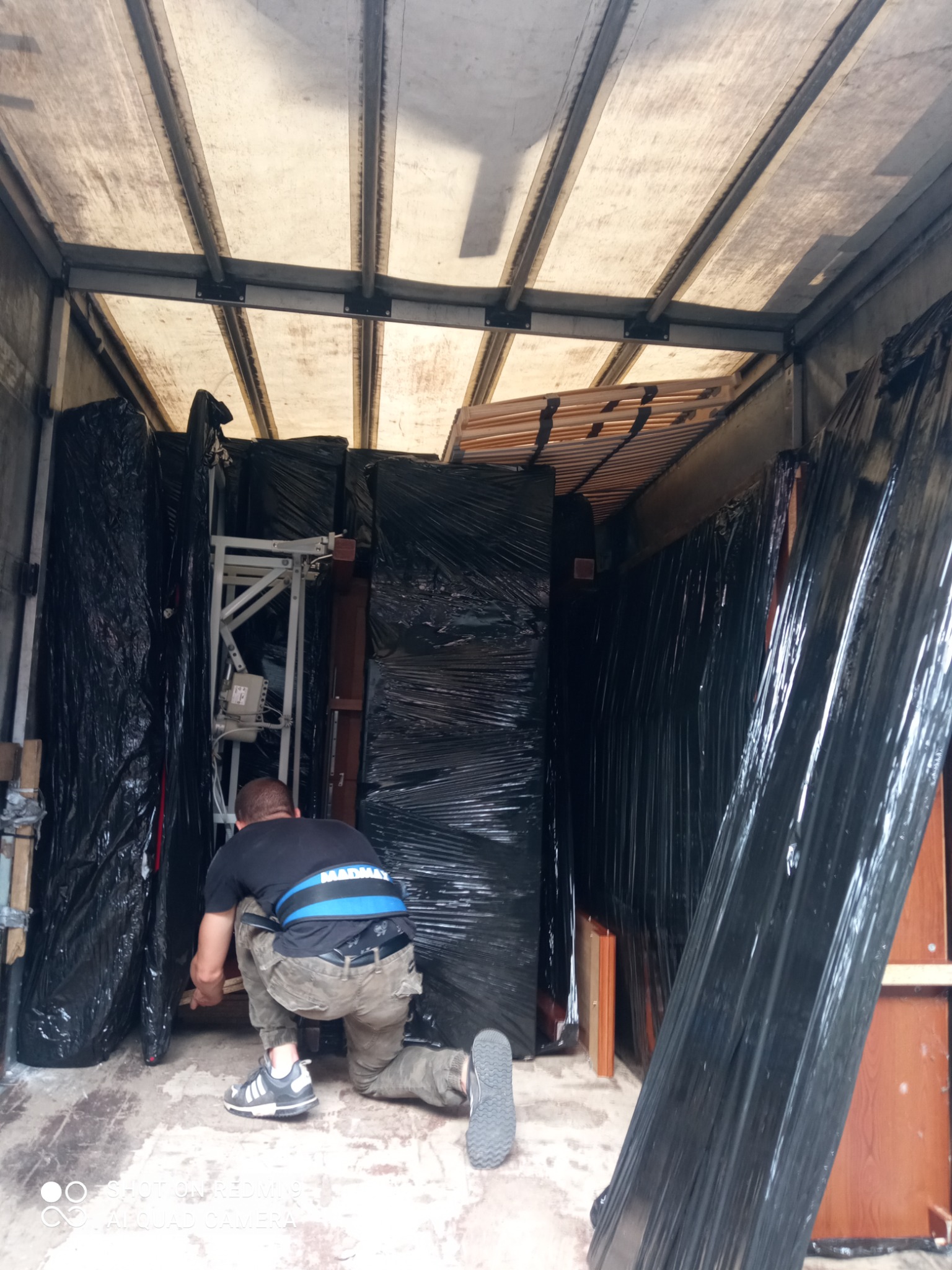 Pracownik w czarnej koszulce i pasie ochronnym na plecy układa meble owinięte czarną folią w przestrzeni ładunkowej pojazdu dostawczego. W tle widoczny stelaż łóżka.