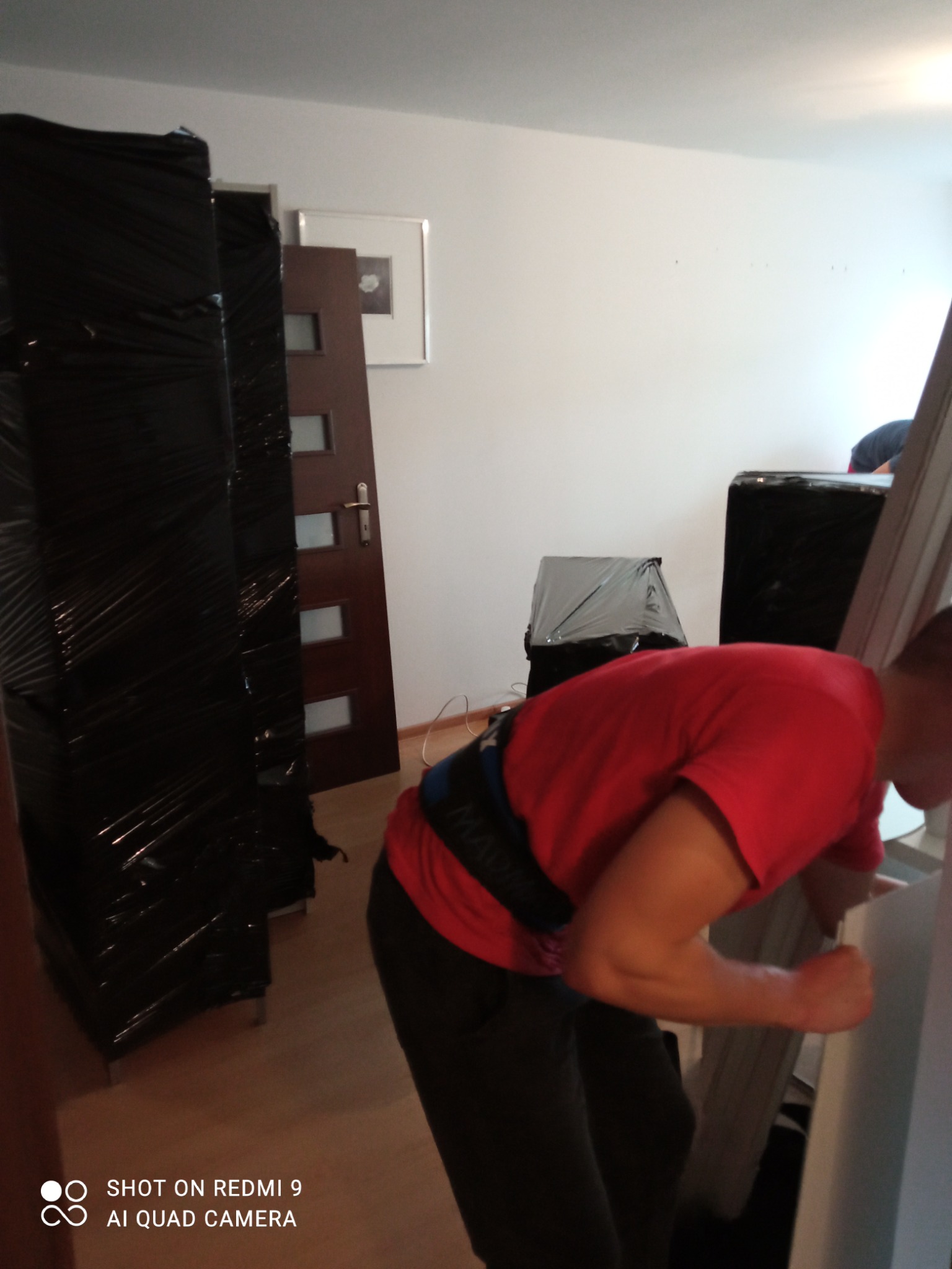 Pracownik w czerwonej koszulce i pasem wspomagającym dźwiga biały mebel podczas przenoszenia zawiniętych w czarną folię mebli w pomieszczeniu.