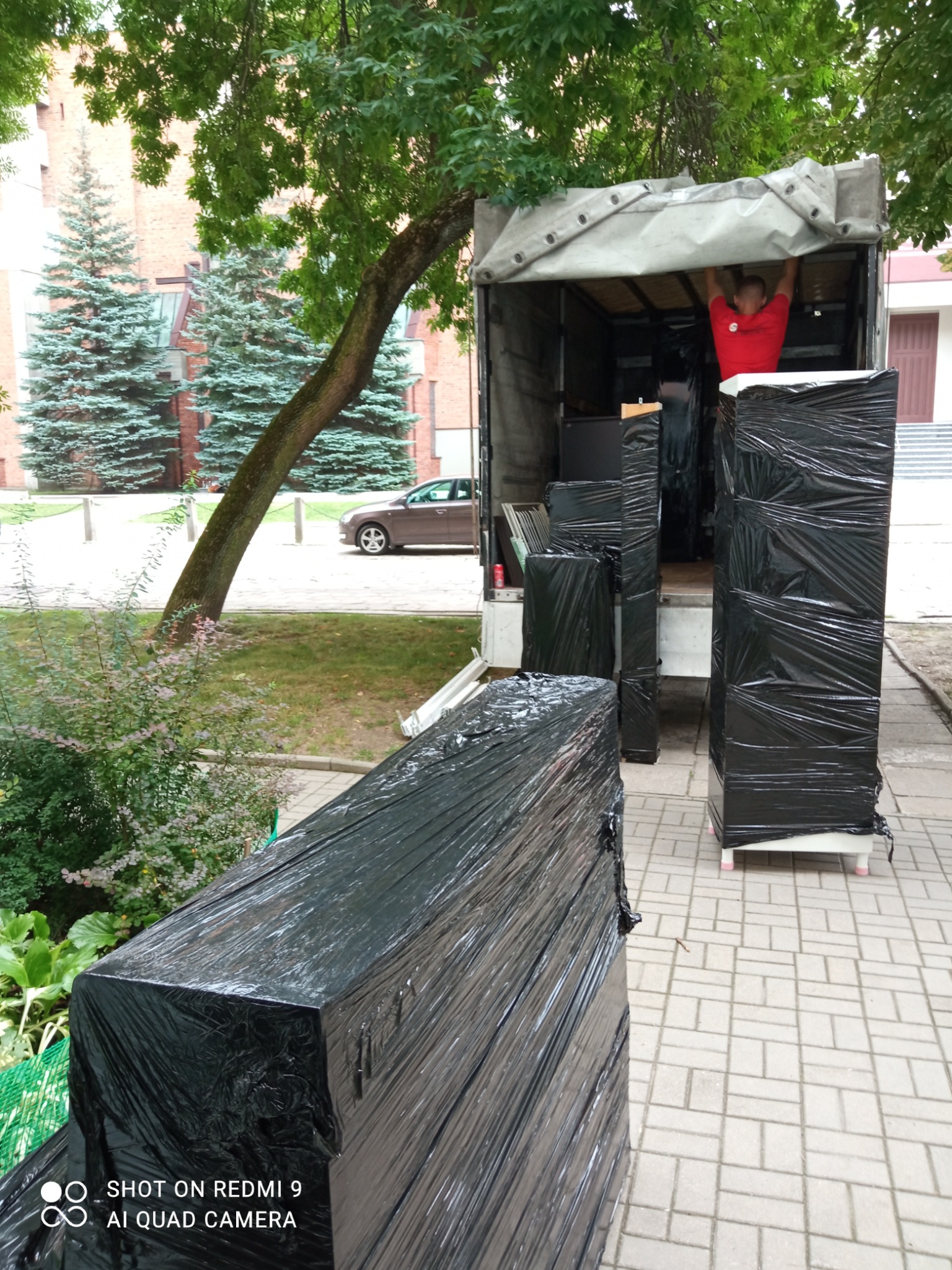 Pracownik w czerwonej koszulce ładuje meble owinięte czarną folią do ciężarówki podczas przeprowadzki; widoczne drzewa i budynek w tle.