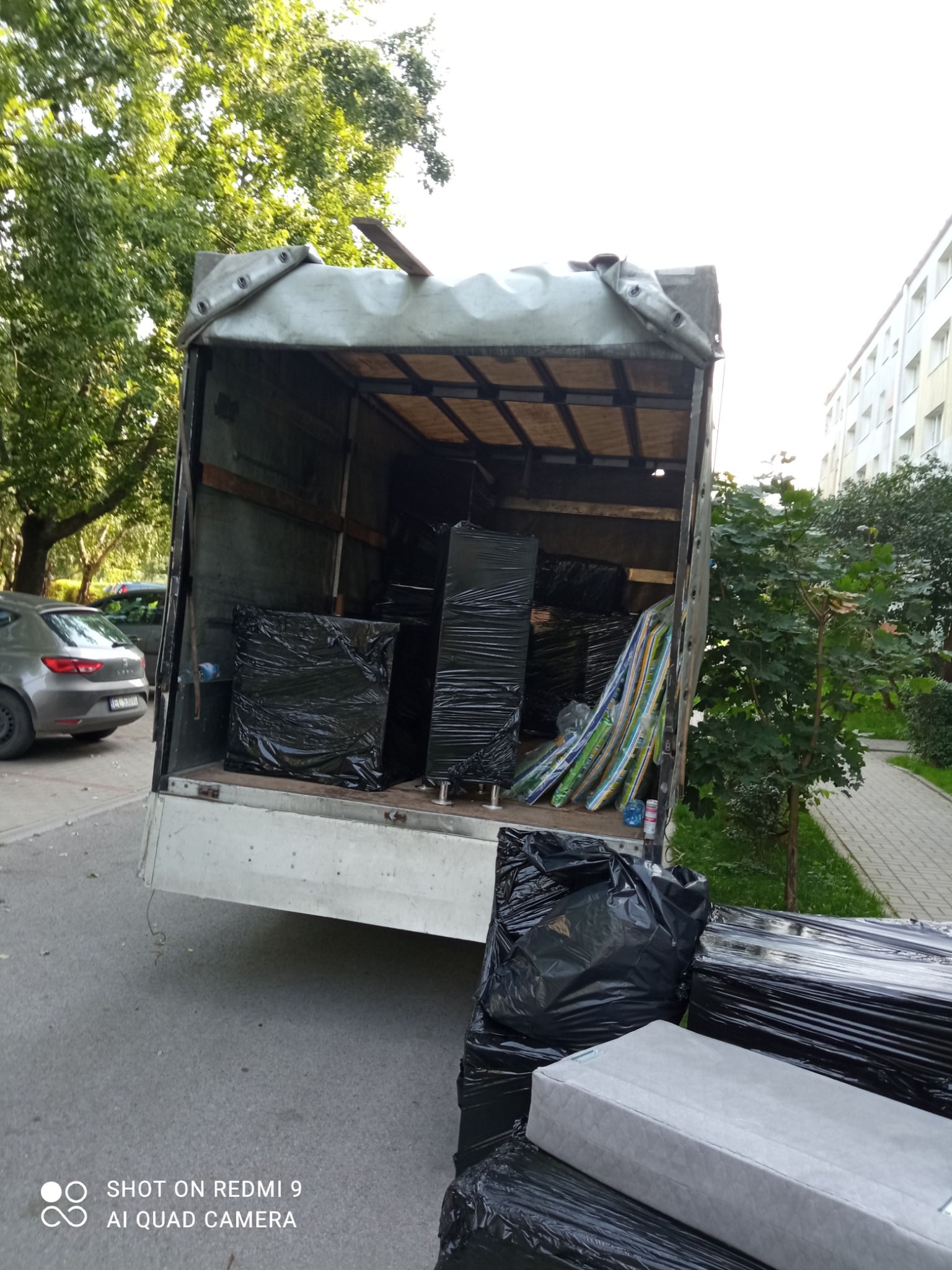 Wnętrze skrzyni ładunkowej samochodu dostawczego wypełnione meblami i przedmiotami zabezpieczonymi czarną folią stretch, widok z zewnątrz na chodnik i zaparkowany samochód osobowy.