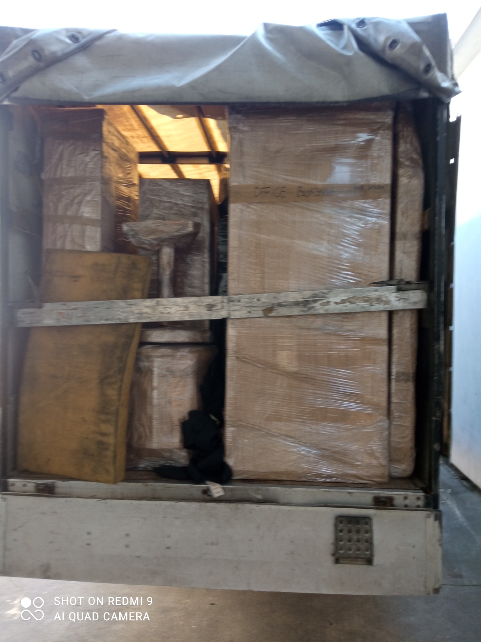 Wnętrze samochodu dostawczego załadowane kartonami i meblami zabezpieczonymi folią stretch, widoczne drewniane deski zabezpieczające ładunek.