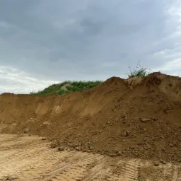 Góra piasku/ziemi z roślinnością na szczycie, ślady opon na piasku, pochmurne niebo.