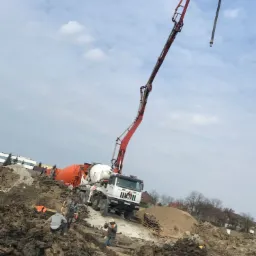 Pompa do betonu na placu budowy, robotnicy przygotowują teren pod fundamenty, widoczne wykopane rowy i błotniste podłoże.