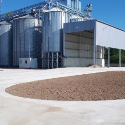 Kompleks silosów zbożowych z rampą załadunkową i przygotowanym terenem pod zagospodarowanie, widoczne dwie ciężarówki na tle błękitnego nieba.