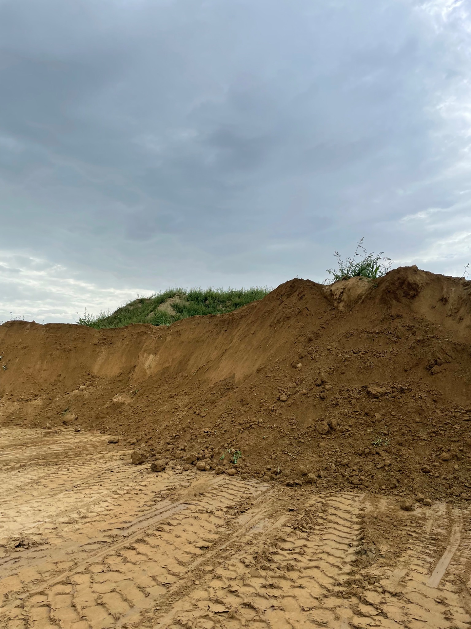 Góra piasku/ziemi z roślinnością na szczycie, ślady opon na piasku, pochmurne niebo.