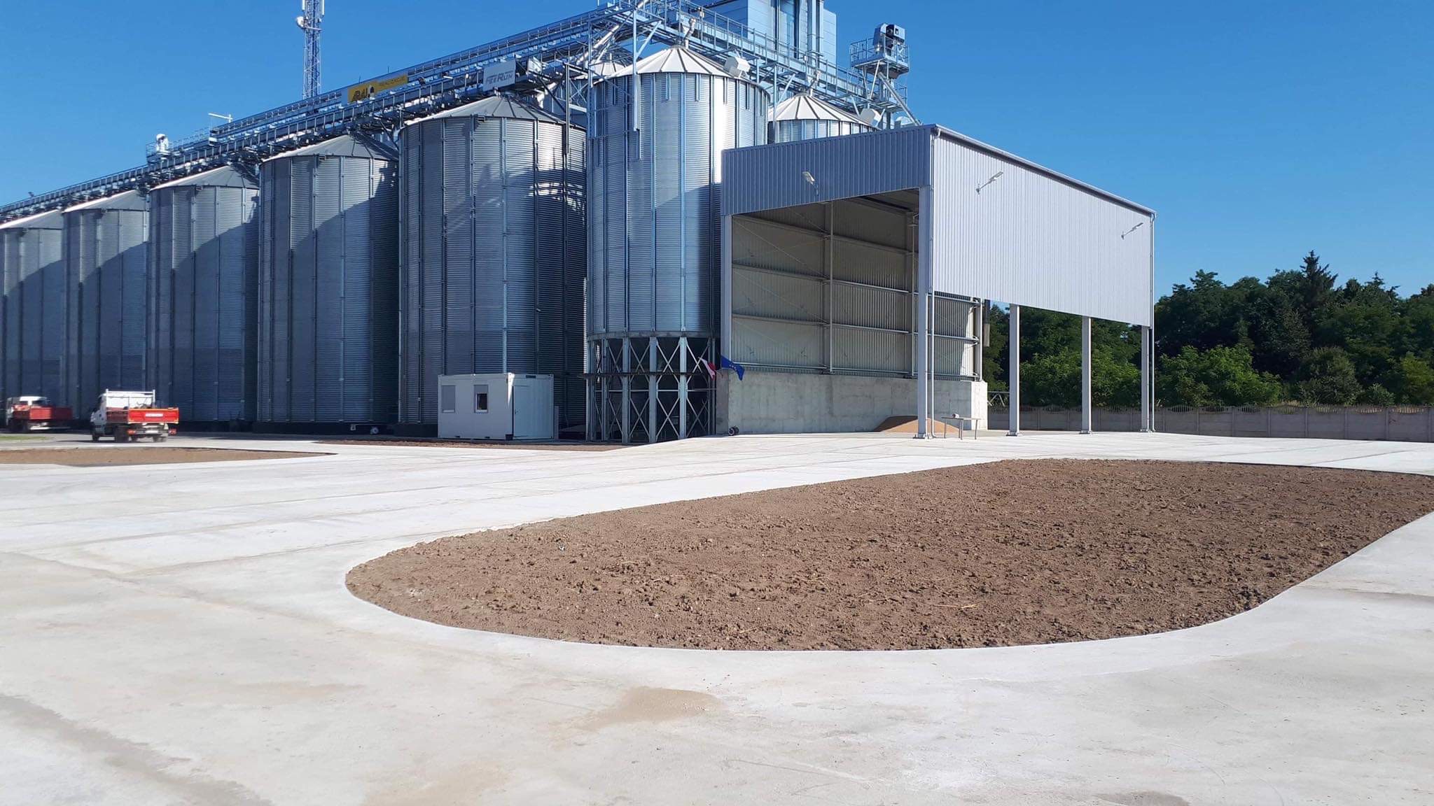 Kompleks silosów zbożowych z rampą załadunkową i przygotowanym terenem pod zagospodarowanie, widoczne dwie ciężarówki na tle błękitnego nieba.