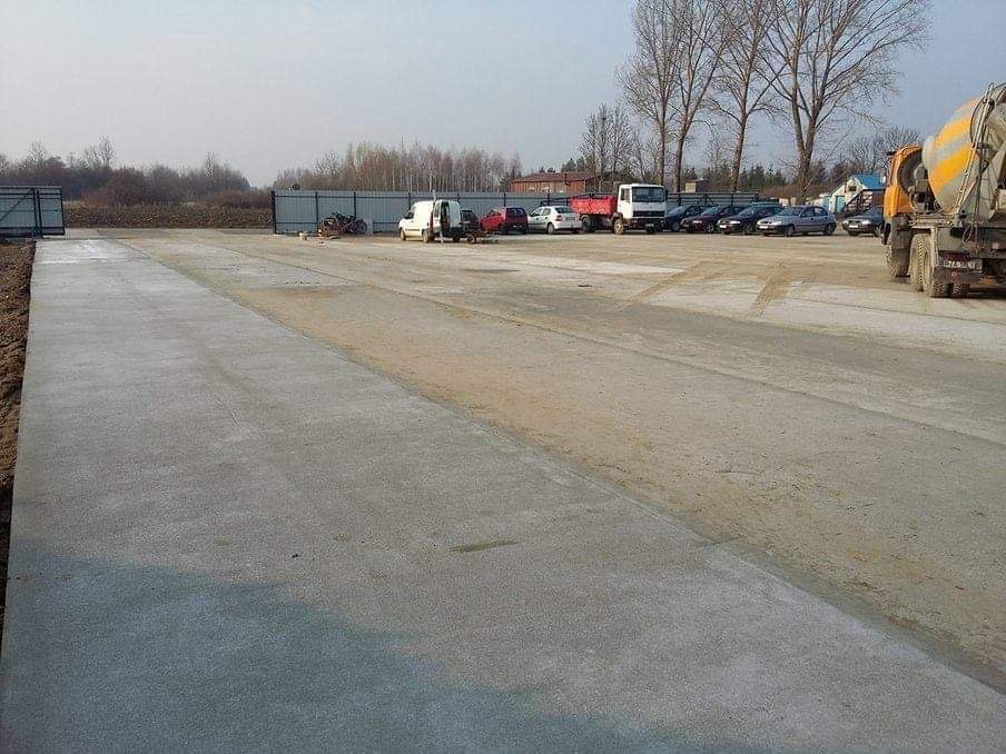 Świeżo wylana, długa, betonowa nawierzchnia placu budowy, z widocznymi śladami opon i zaparkowanymi pojazdami w tle, w tym betoniarka i samochody osobowe, całość otoczona metalowym ogrodzeniem.