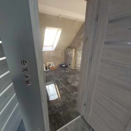 PRO-BUD Przemysław Wrzesniewski - Wnętrze łazienki na poddaszu w trakcie remontu, widok przez otwarte drzwi, z widocznymi czarnymi płytkami na podłodze, oknem dachowym i instalacją elektryczną w ścianie.