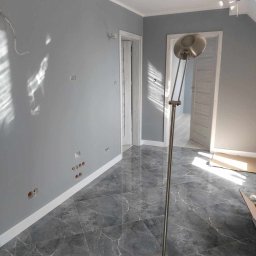 PRO-BUD Przemysław Wrzesniewski - Świeżo pomalowane szare ściany w pokoju z marmurową podłogą, widoczne puszki elektryczne bez osprzętu i lampa podłogowa.