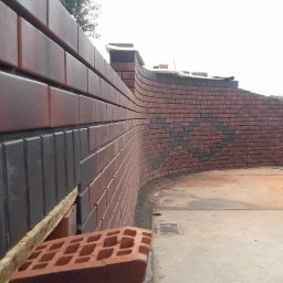 PRO-BUD Przemysław Wrzesniewski - Budowa zakrzywionego muru z cegły klinkierowej, widoczny fragment nowej, czarnej elewacji oraz dalsza część ściany z czerwonej cegły, w tle zieleń drzew.