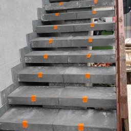 PRO-BUD Przemysław Wrzesniewski - Montaż szarych płytek na schodach zewnętrznych z użyciem pomarańczowych krzyżyków dystansowych, widoczny pracownik w odblaskowej kamizelce i wiadro z klejem.