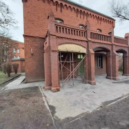 PRO-BUD Przemysław Wrzesniewski - Ceglany budynek z charakterystycznym portykiem w trakcie renowacji, z widocznymi rusztowaniami i fragmentami odkrytej elewacji.