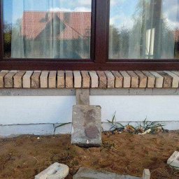 PRO-BUD Przemysław Wrzesniewski - Fragment elewacji domu z oknem, parapet wykończony cegłą klinkierową, podpora z betonowego bloczka w ziemi, widoczne ślady prac budowlanych i niedokończone tynkowanie.
