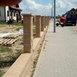 PRO-BUD Przemysław Wrzesniewski - Wznoszony mur z prefabrykatów betonowych, widoczne palety z materiałem, betoniarka na chodniku, domy w tle.