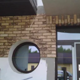 Fragment elewacji domu z okrągłym oknem obłożony dekoracyjną cegłą w odcieniach beżu i brązu, widoczne prace wykończeniowe.