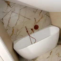 Nowoczesna biała wanna wolnostojąca z miedzianą armaturą na tle płytek imitujących marmur w łazience na poddaszu.