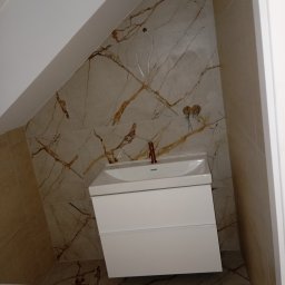 PRO-BUD Przemysław Wrzesniewski - Ukośna łazienka na poddaszu z marmurowymi płytkami, białą szafką pod umywalkę i miedzianą baterią; widoczne niedokończone podłączenia elektryczne.