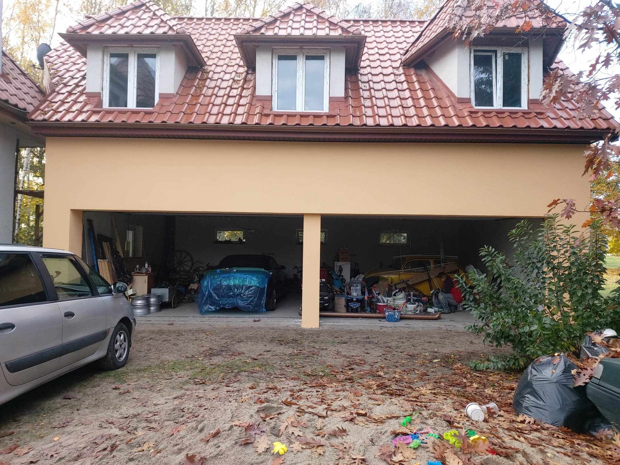 Dwukondygnacyjny budynek z garażem na parterze, elewacja w odcieniach beżu i brązu, czerwona dachówka, na podwórku porozrzucane liście i zabawki, w garażu widoczne dwa samochody przykryte folią...