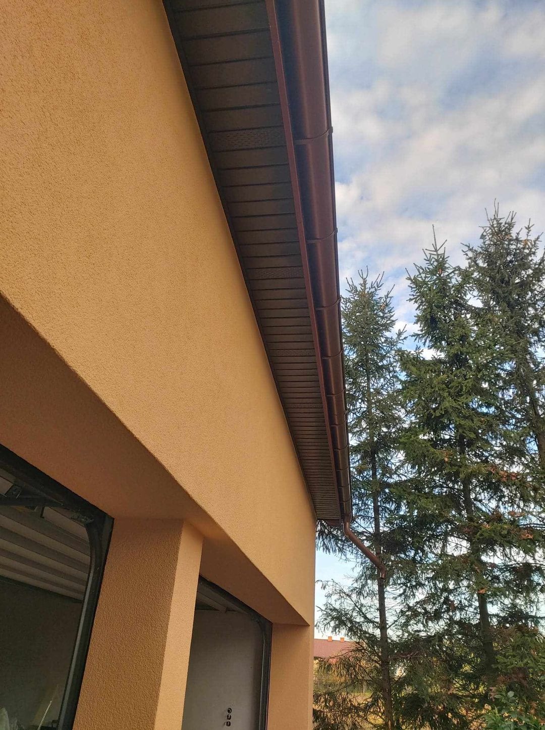 Jasnożółta elewacja domu z brązowymi rynnami, widok z dołu na tle błękitnego nieba i zielonych drzew iglastych.