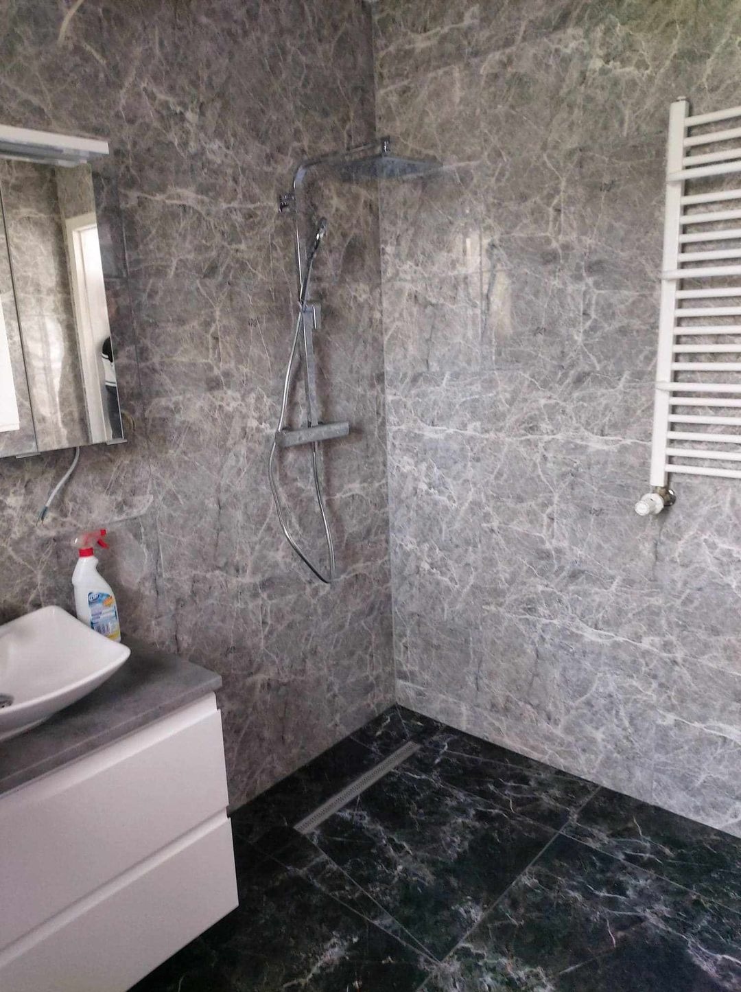 Wykończona łazienka z szarymi marmurkowymi płytkami na ścianach, czarną marmurkową podłogą z odpływem liniowym, białą szafką z umywalką i grzejnikiem drabinkowym.