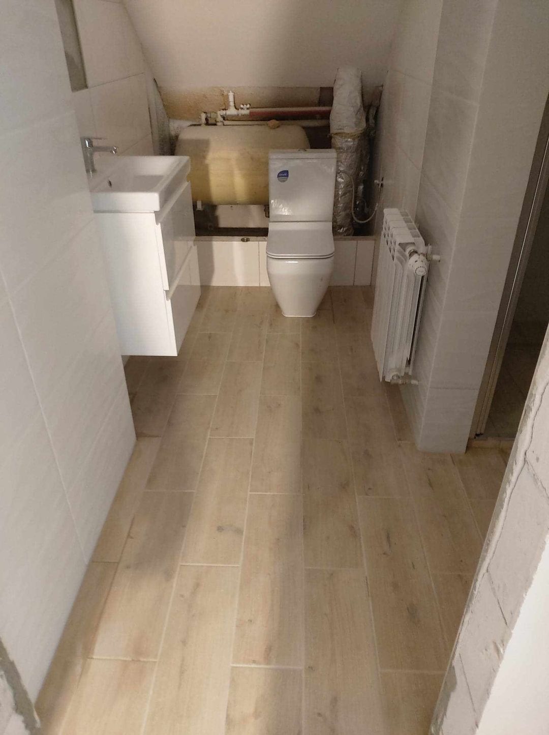 Wnętrze łazienki w trakcie remontu: białe płytki ścienne, beżowe płytki podłogowe imitujące drewno, biała umywalka z szafką, toaleta kompaktowa oraz grzejnik żeberkowy. Widoczny bojler i rury...
