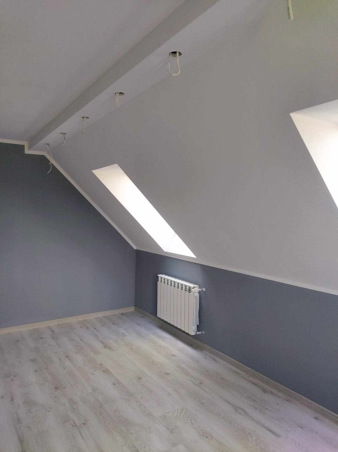 Pokój na poddaszu po remoncie: szare ściany, biały grzejnik, jasne panele podłogowe, okno dachowe i otwory na oświetlenie sufitowe z wystającymi kablami.