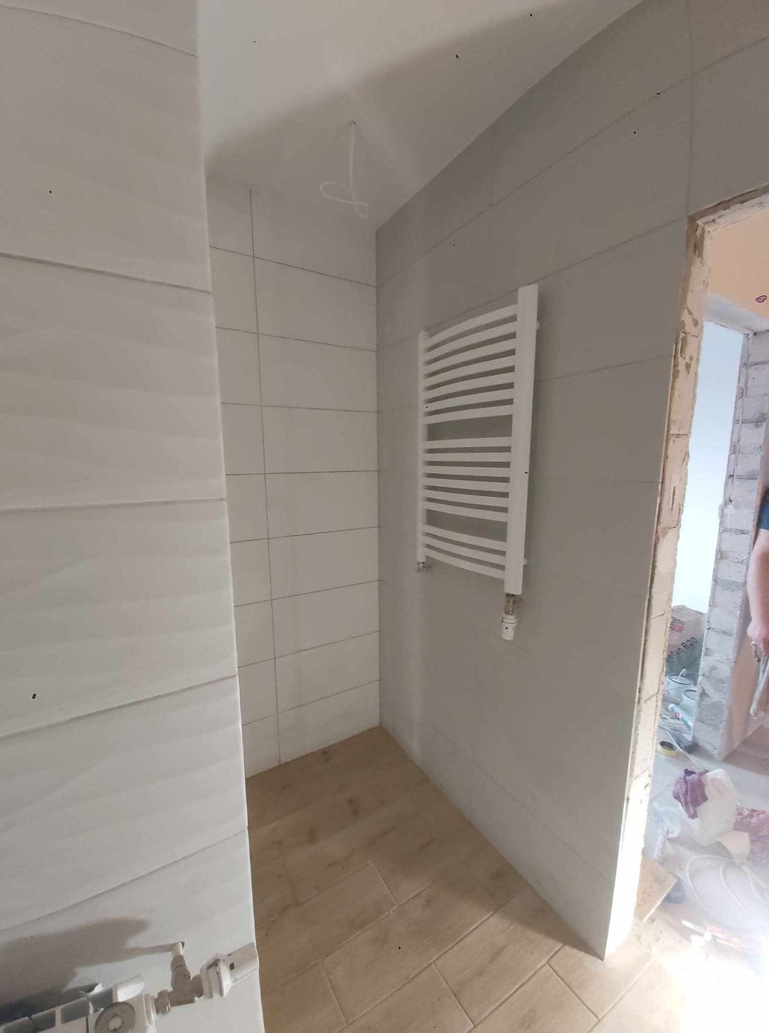 Wykończona łazienka z ułożonymi płytkami imitującymi drewno na podłodze, szare płytki na ścianach i zamontowanym białym grzejnikiem drabinkowym; widoczne niedokończone obramowanie drzwi.