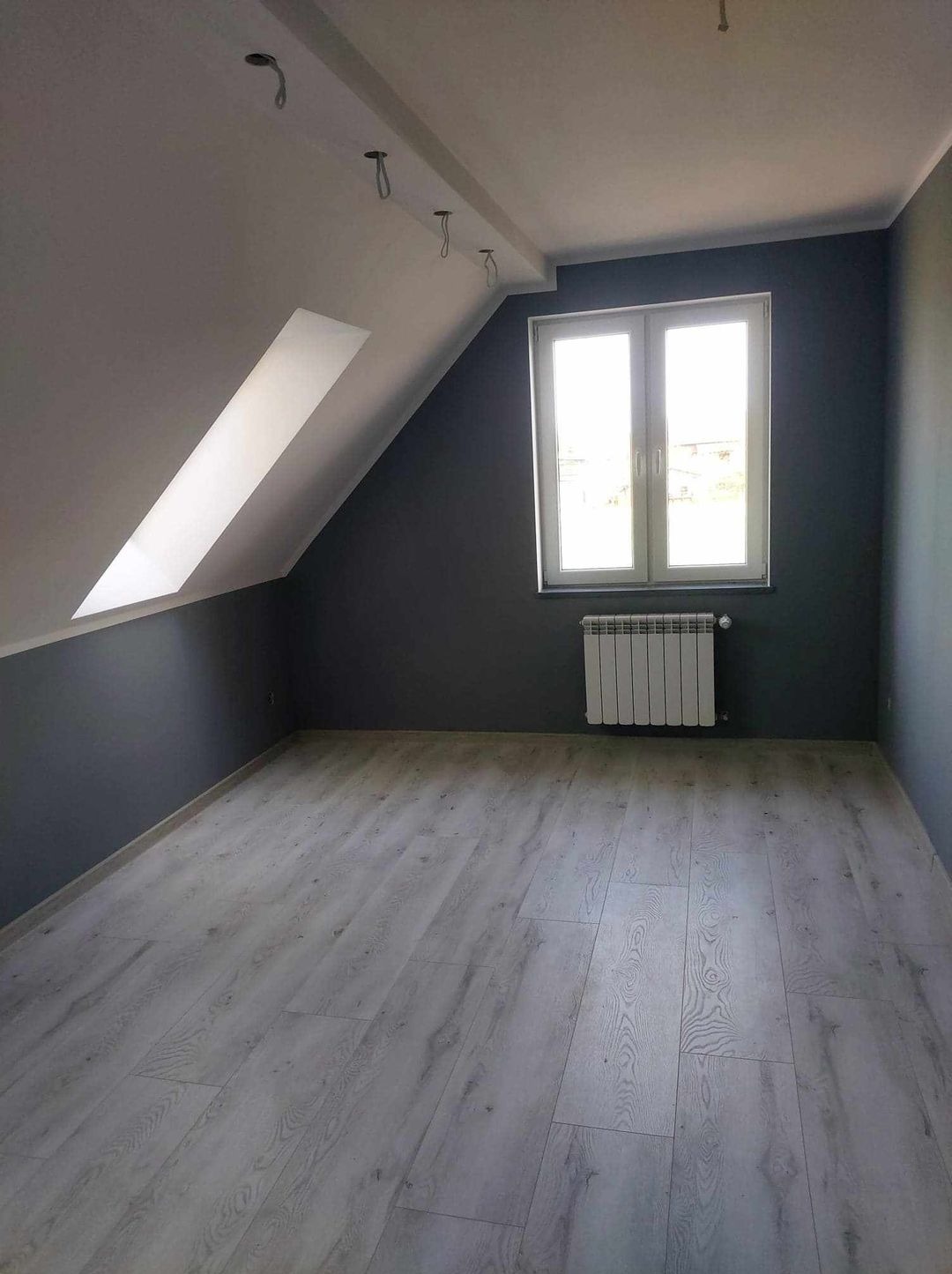 Poddasze po remoncie: szare ściany, jasne panele podłogowe, białe okno z grzejnikiem, widoczne kable od oświetlenia w suficie.