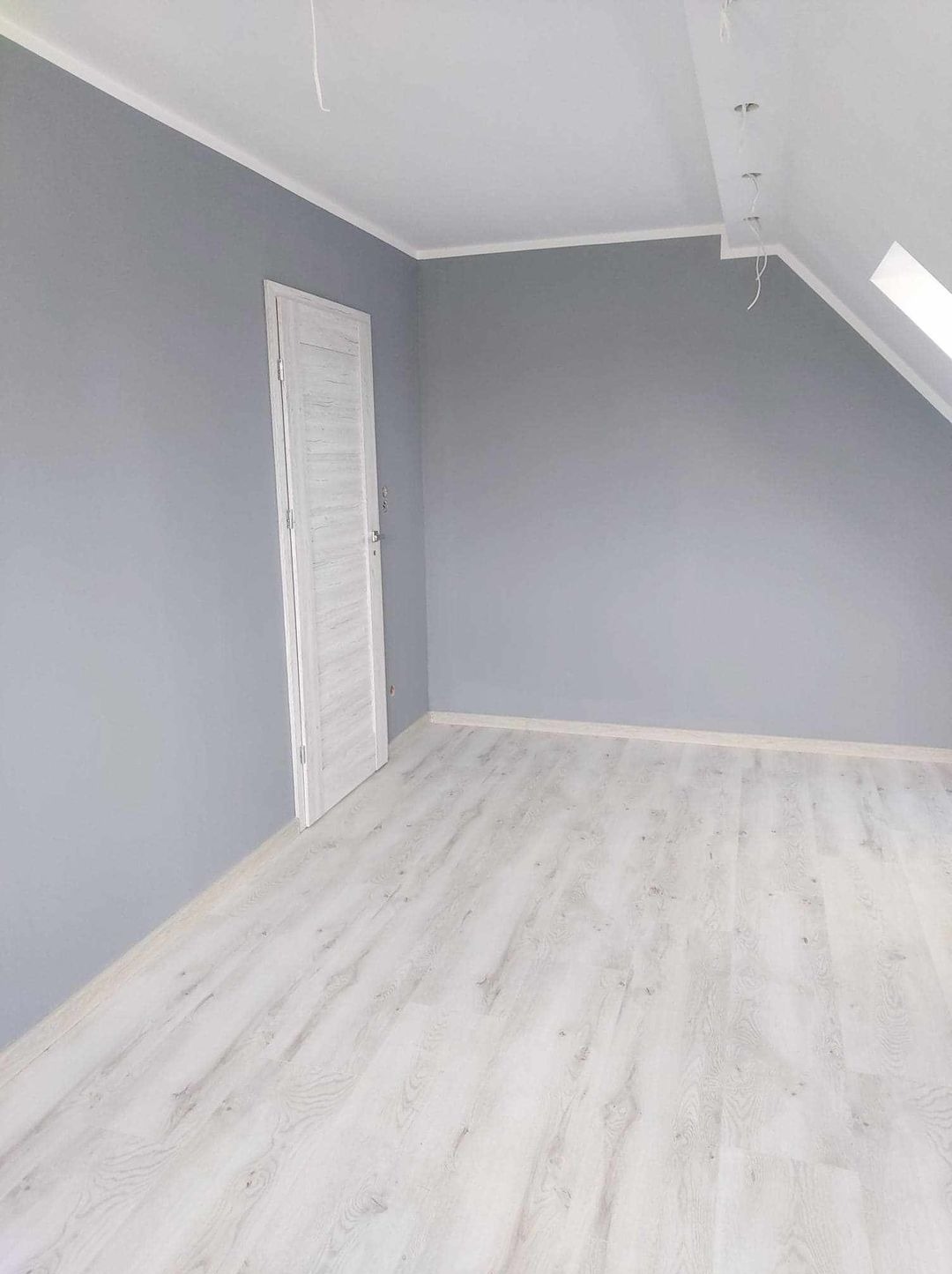 Pokój na poddaszu po remoncie: szare ściany, białe panele podłogowe z widoczną strukturą drewna, białe drzwi z delikatnym wzorem i skośny sufit z oknem dachowym.