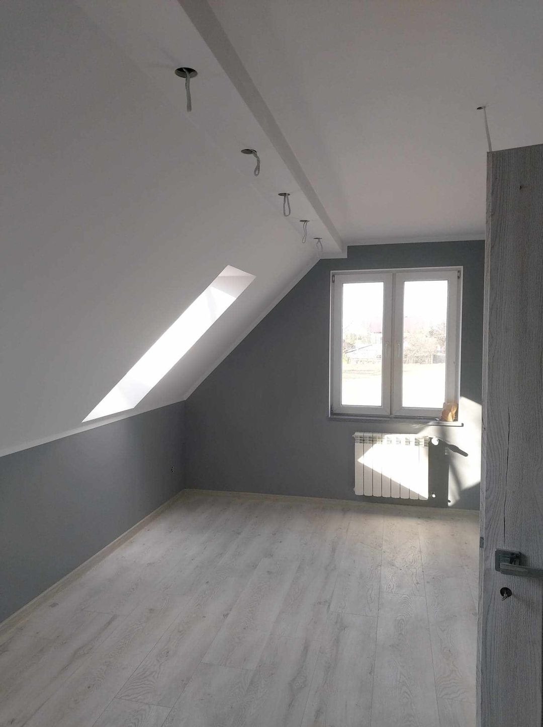 Pokój na poddaszu po remoncie: szare ściany, biały sufit, jasne panele podłogowe, okno dachowe i grzejnik pod oknem, widoczne kable od oświetlenia sufitowego.