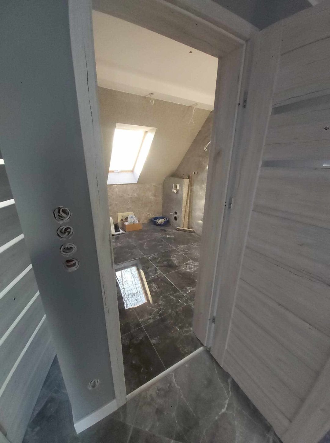 Wnętrze łazienki na poddaszu w trakcie remontu, widok przez otwarte drzwi, z widocznymi czarnymi płytkami na podłodze, oknem dachowym i instalacją elektryczną w ścianie.