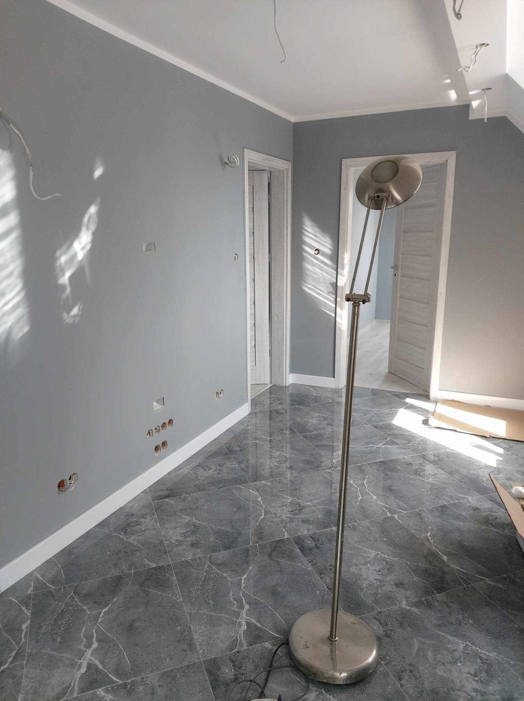 Świeżo pomalowane szare ściany w pokoju z marmurową podłogą, widoczne puszki elektryczne bez osprzętu i lampa podłogowa.