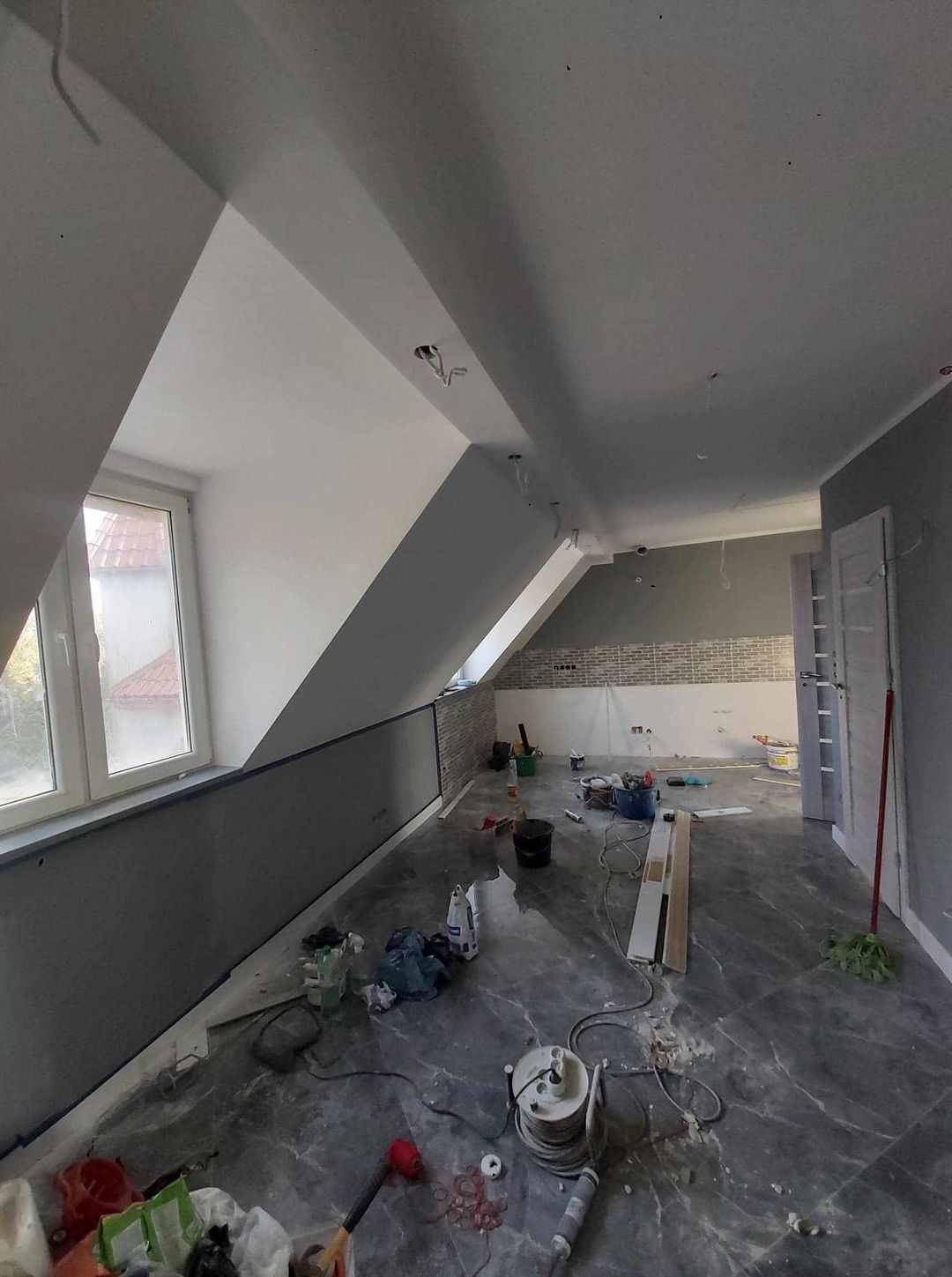 Pokój na poddaszu w trakcie prac wykończeniowych: świeżo pomalowane ściany w odcieniach bieli i szarości, widoczne narzędzia malarskie, kable wystające z sufitu oraz fragment ściany wyłożonej cegłą.