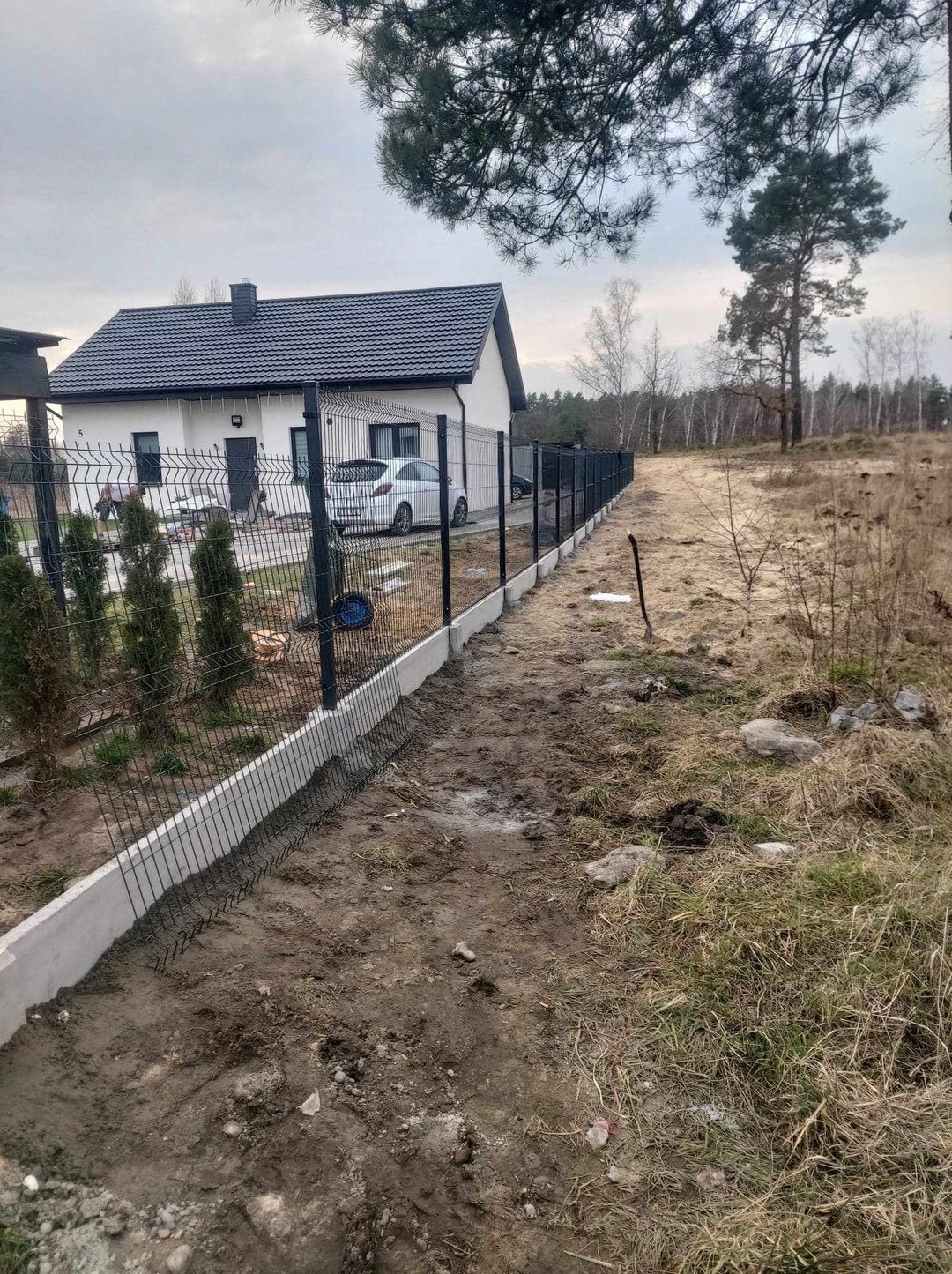 Montaż ogrodzenia panelowego z podmurówką betonową przy nowym domu jednorodzinnym, widoczne elementy mocujące siatkę do podmurówki, teren wokół częściowo uprzątnięty, w tle drzewa i biały samochód.