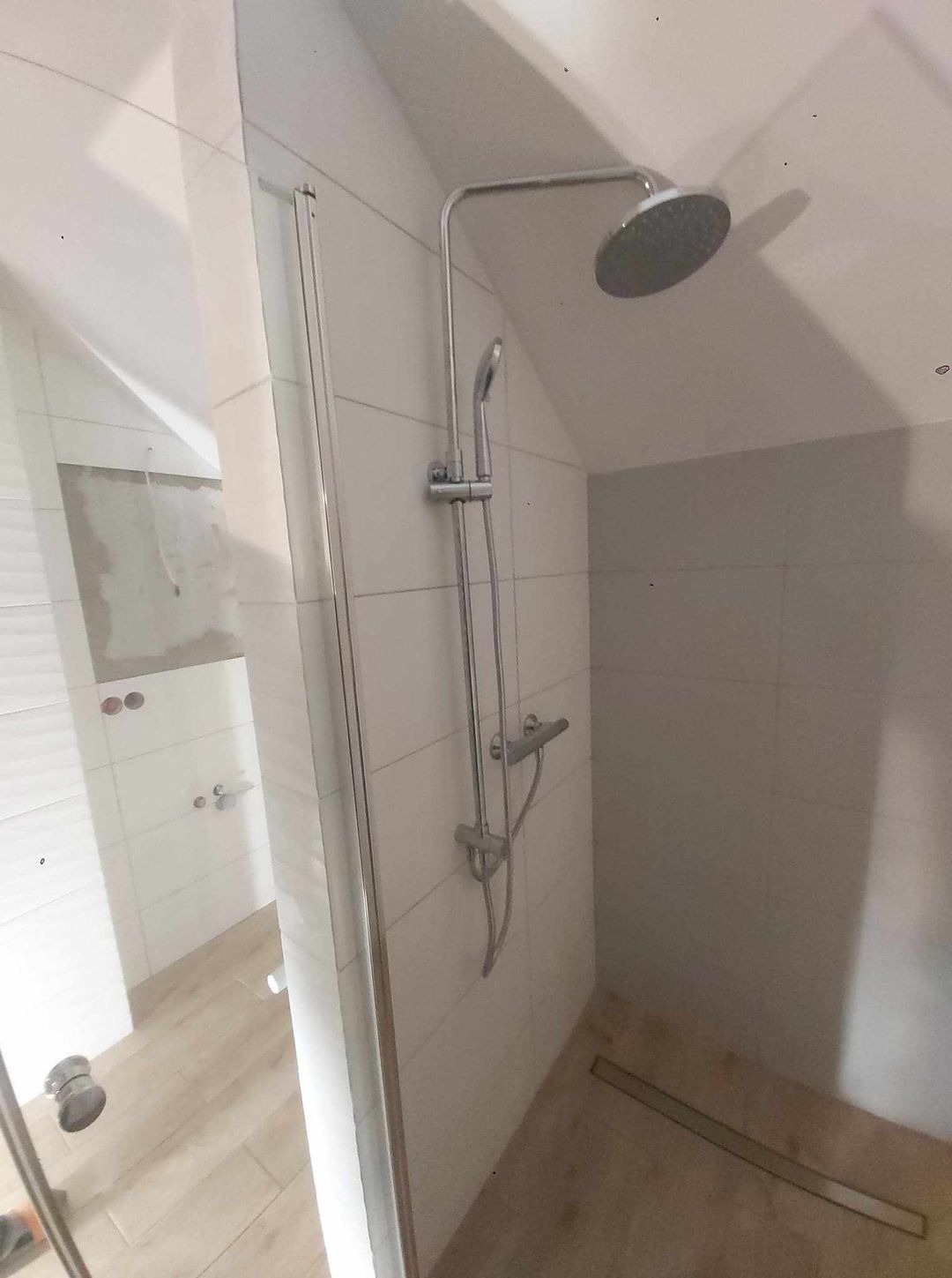 Nowoczesny prysznic z deszczownicą i baterią termostatyczną w jasnej łazience z białymi płytkami i odpływem liniowym w podłodze, widok na częściowo wykończone wnętrze z przygotowanymi otworami...