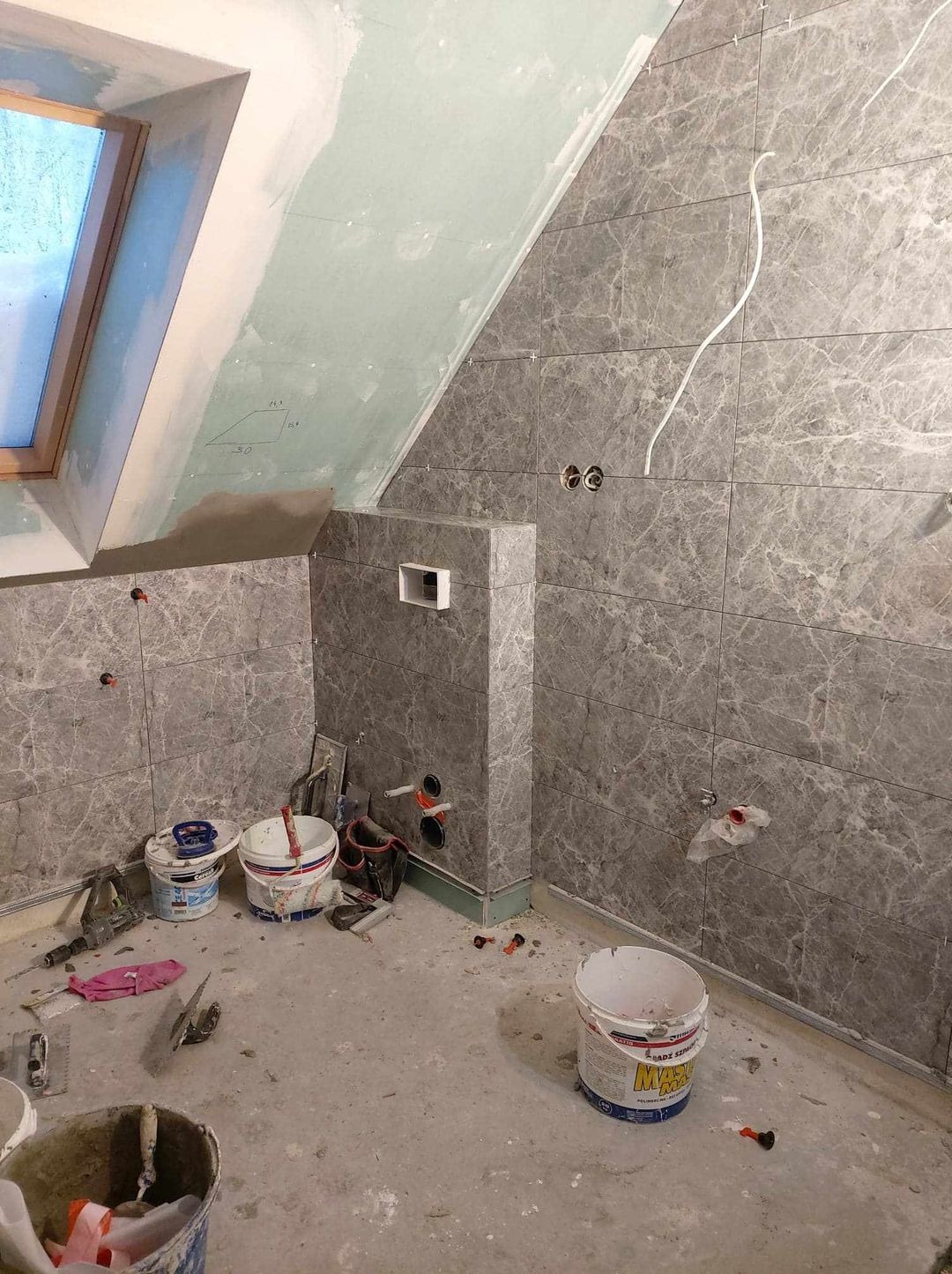 Remont łazienki na poddaszu: szare marmurowe płytki na ścianach i zabudowie, widoczne narzędzia i materiały budowlane, okno dachowe, instalacja elektryczna w trakcie montażu.