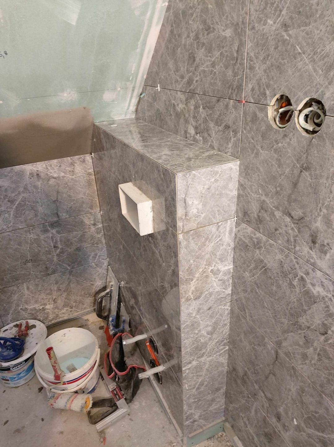 Ściana łazienki w trakcie wykańczania szarymi płytkami imitującymi marmur, widoczne puszki elektryczne, narzędzia i wiaderka z zaprawą na podłodze.