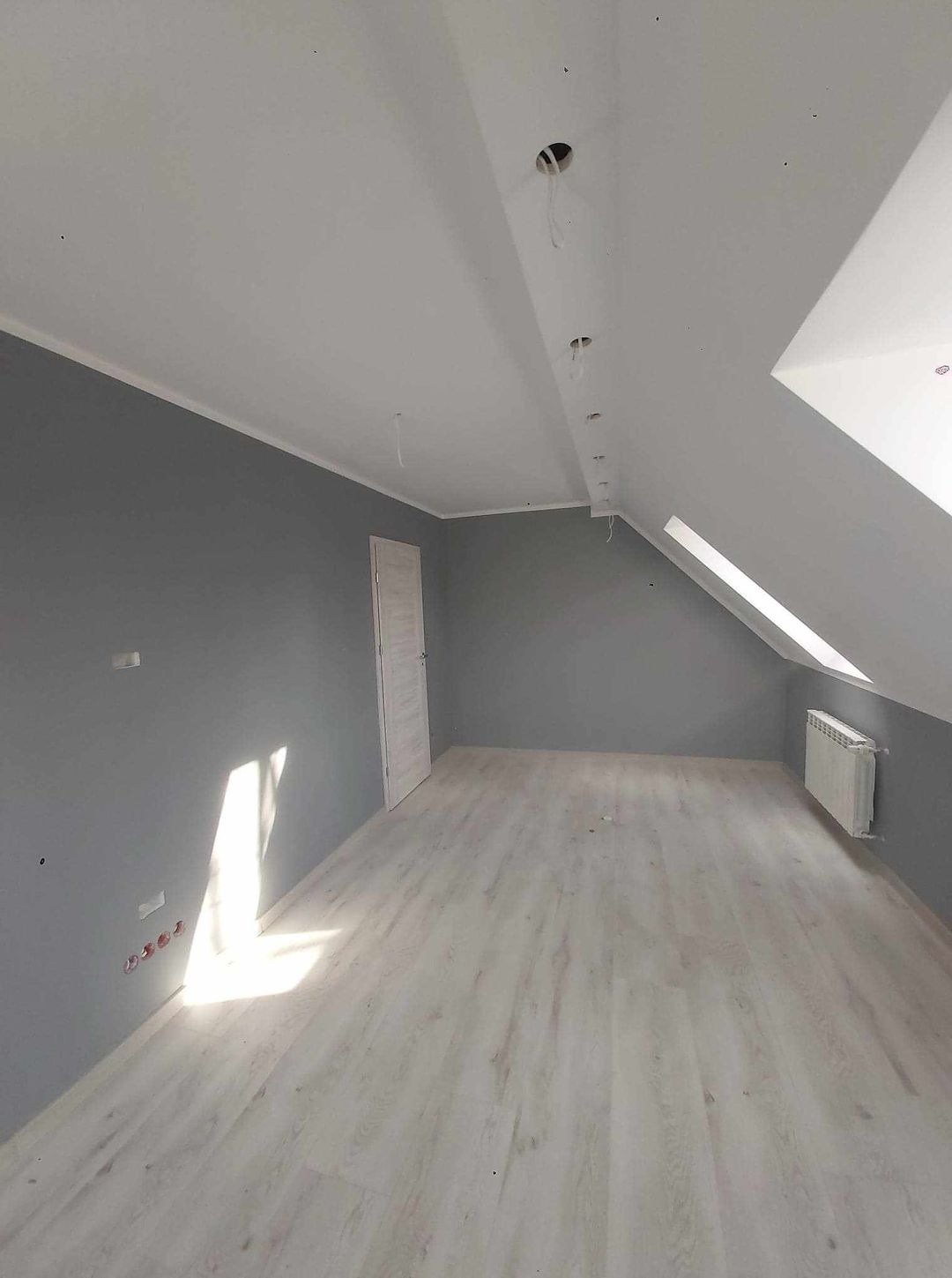 Poddasze po remoncie: szare ściany, jasna podłoga z paneli, białe drzwi, grzejnik pod oknem dachowym, przygotowane punkty oświetleniowe w suficie.