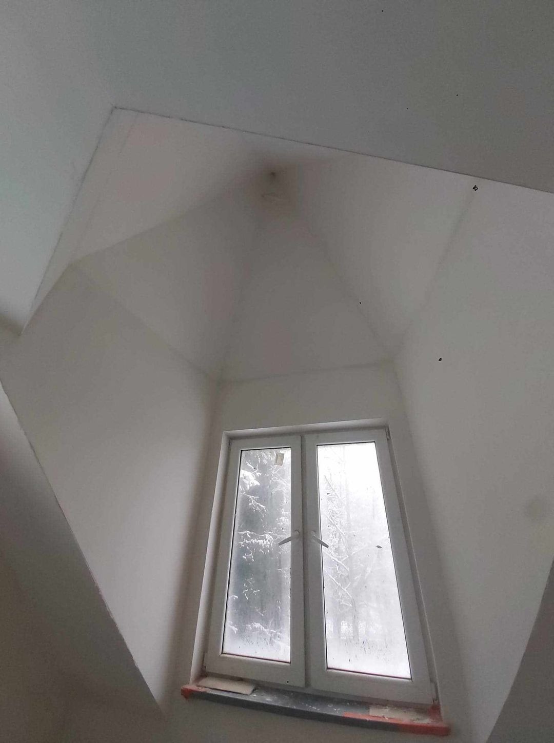 Wnętrze pokoju na poddaszu z oknem, ściany i sufit świeżo pomalowane na biało, widoczne ślady po niedoskonałościach na suficie, za oknem zima.