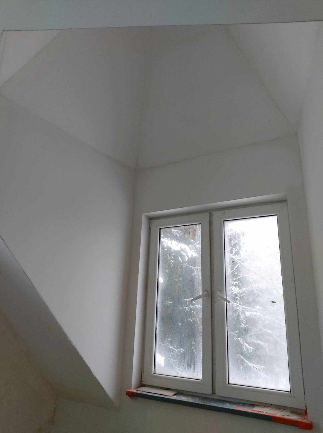 Świeżo pomalowane na biało wnętrze pokoju z oknem, widoczne ślady po taśmie malarskiej na parapecie, z zewnątrz widok zaśnieżonych drzew.