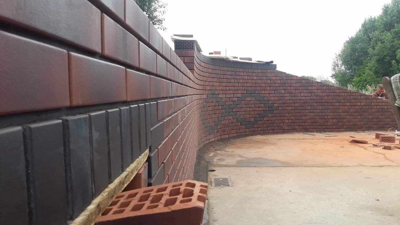 Budowa zakrzywionego muru z cegły klinkierowej, widoczny fragment nowej, czarnej elewacji oraz dalsza część ściany z czerwonej cegły, w tle zieleń drzew.